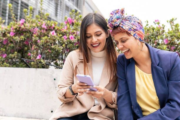 Two women sitting outside smiling as they view a phone screen together